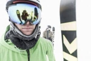 Gros plan sur un skieur portant un masque de ski reflétant la montagne, avec un second skieur en arrière-plan dans une ambiance hivernale.